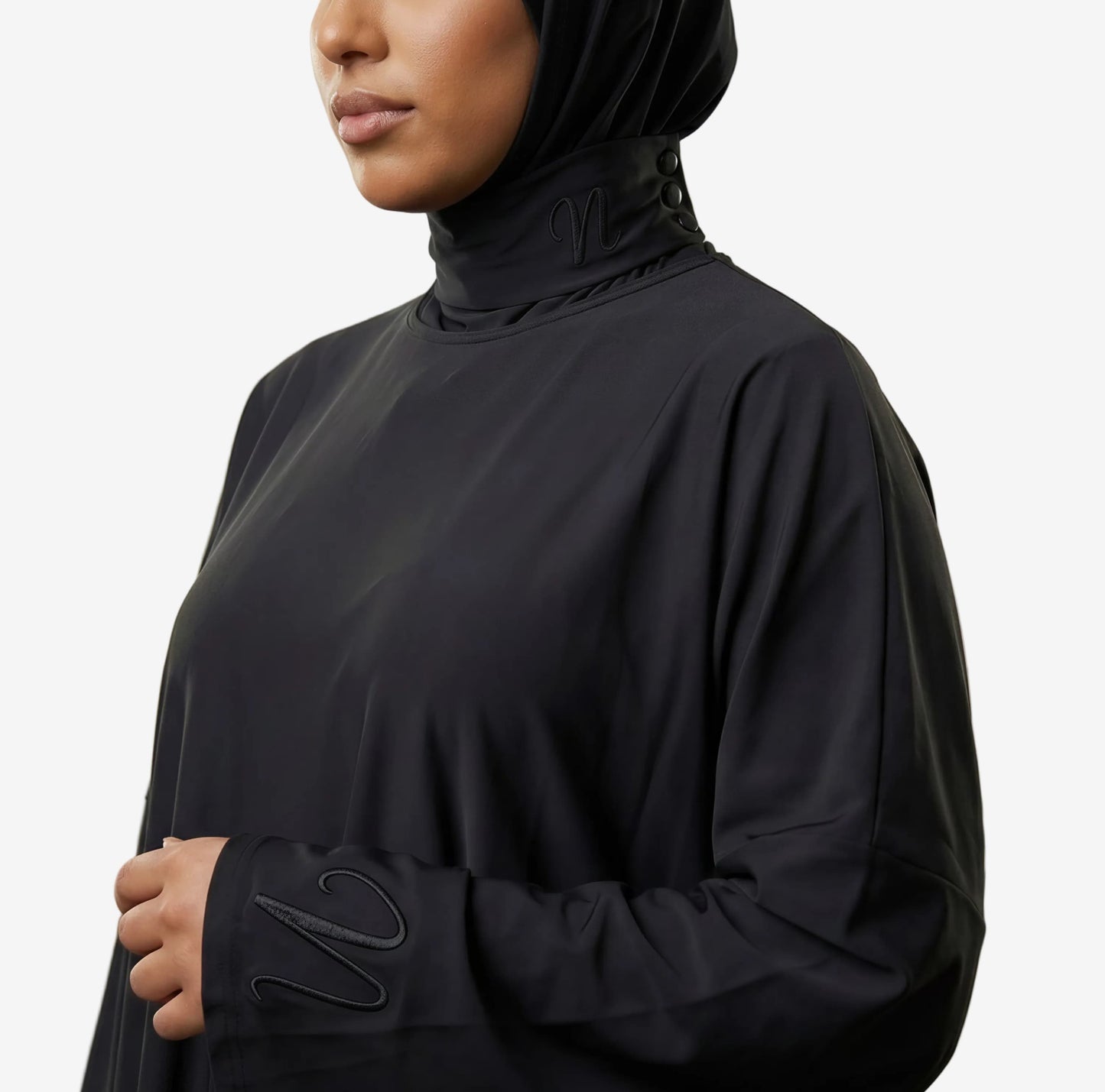 Femme portant un burkini 3 pièces noir avec cagoule intégrée - coupe ample et confortable - zoom sur les détails de la cagoule et du tissu ainsi que le logo